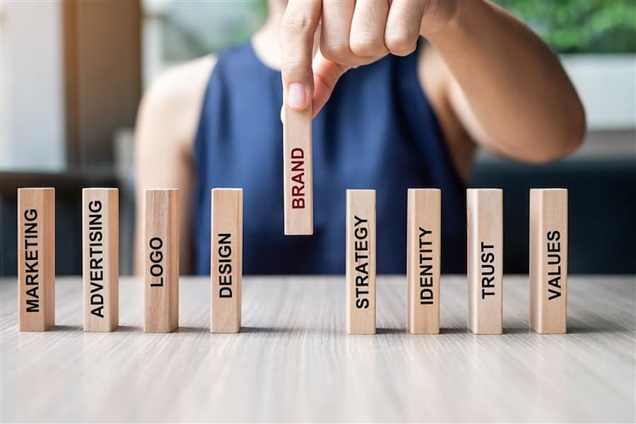 A person placing a wooden block labeled 'BRAND' among a row of standing blocks reading Marketing, Advertising, Logo, Design, Strategy, Identity, Trust, and Values — illustrating the key components of What Is Branding Strategy in Marketing